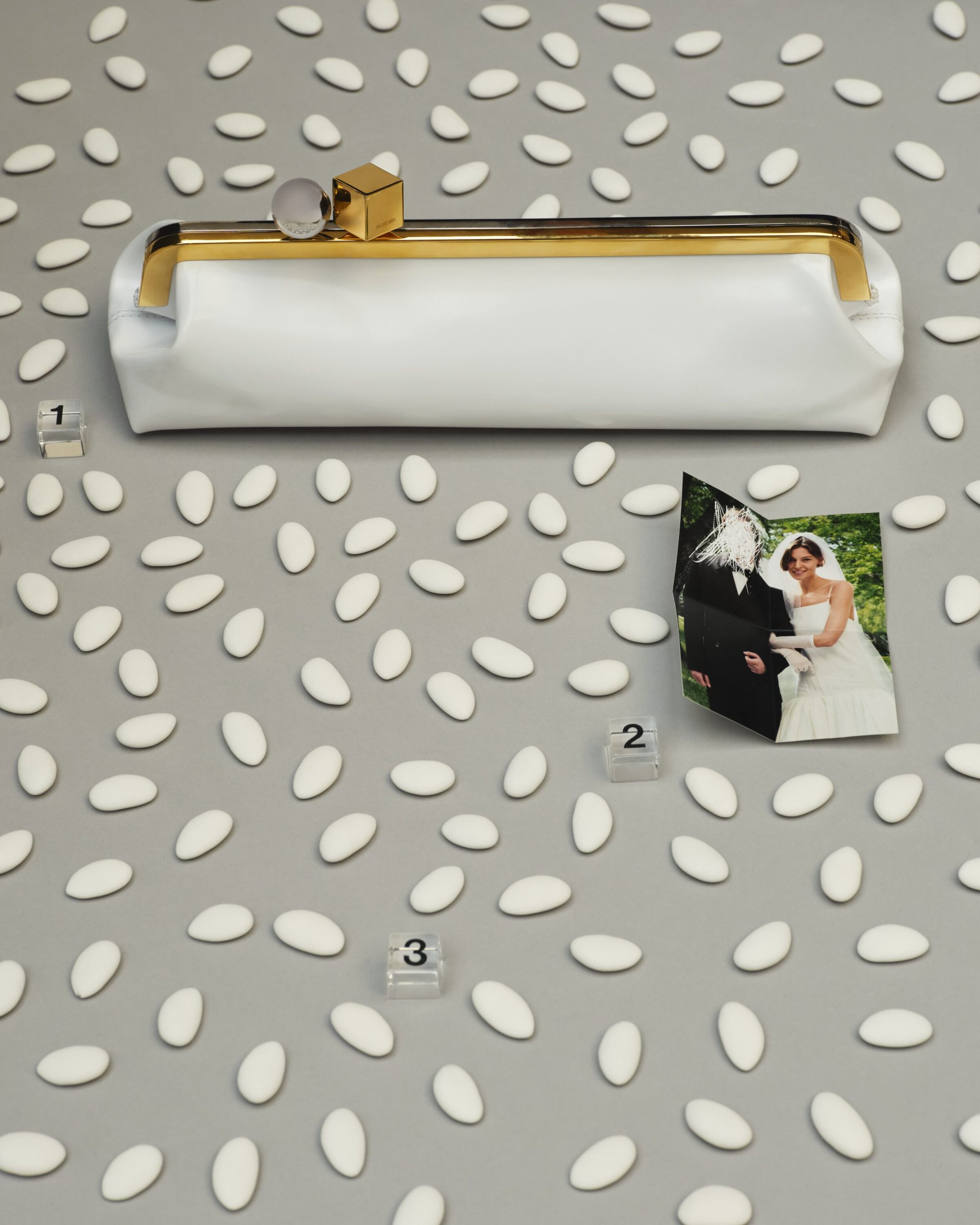 White leather clutch with gold frame and geometric charms on a grey surface scattered with white Jordan almonds and a wedding photo.