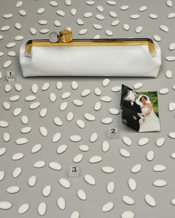 White leather clutch with gold frame and geometric charms on a grey surface scattered with white Jordan almonds and a wedding photo.