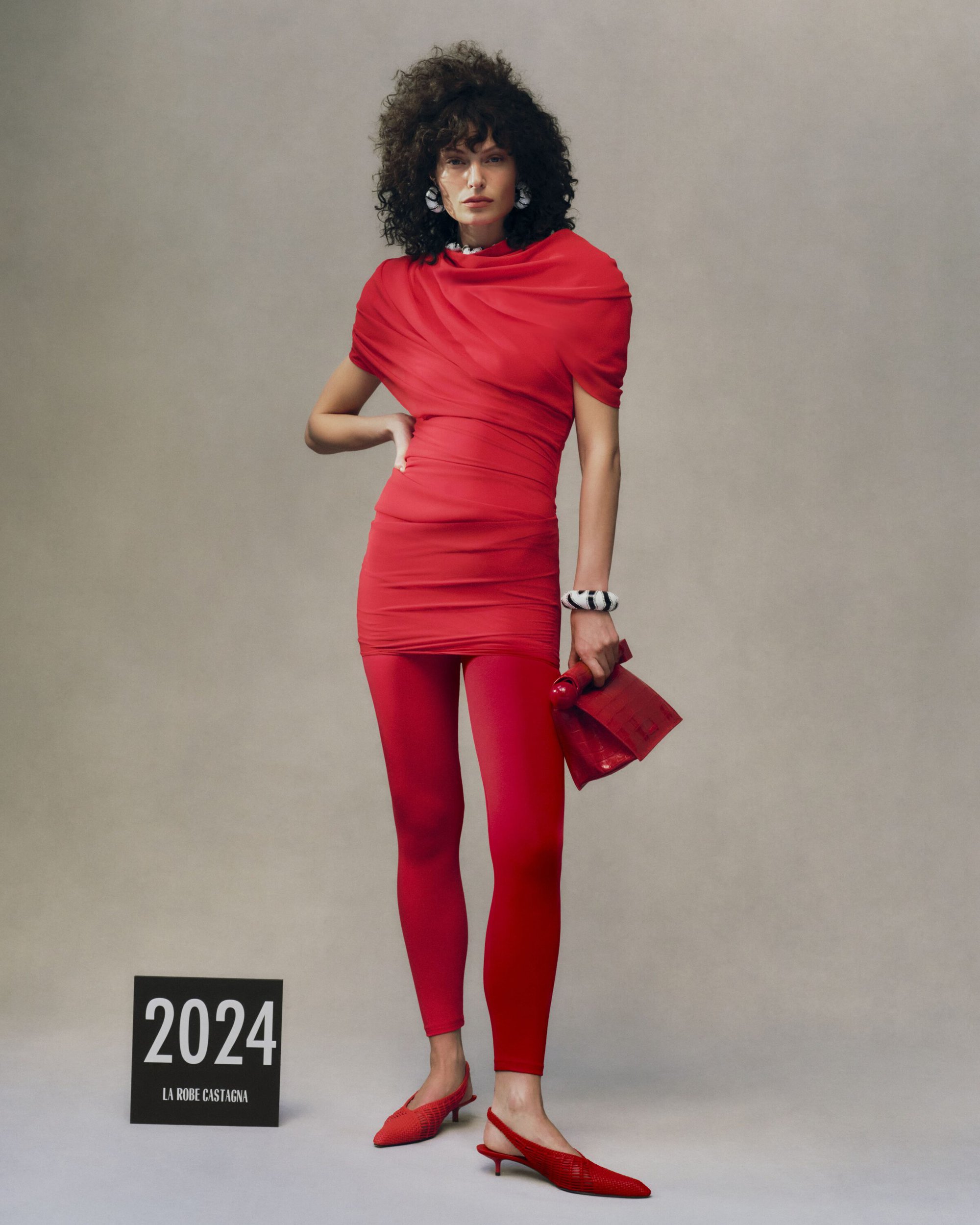 Model in a red dress and leggings holding a matching red handbag against a neutral grey studio background