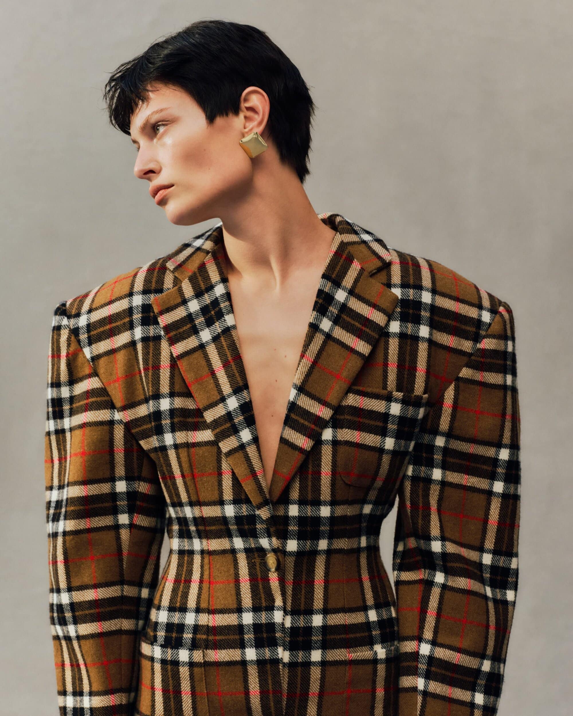 Model with dark short hair wearing a plaid blazer and square gold earrings against a grey textured studio background.