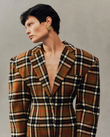 Model with dark short hair wearing a plaid blazer and square gold earrings against a grey textured studio background.