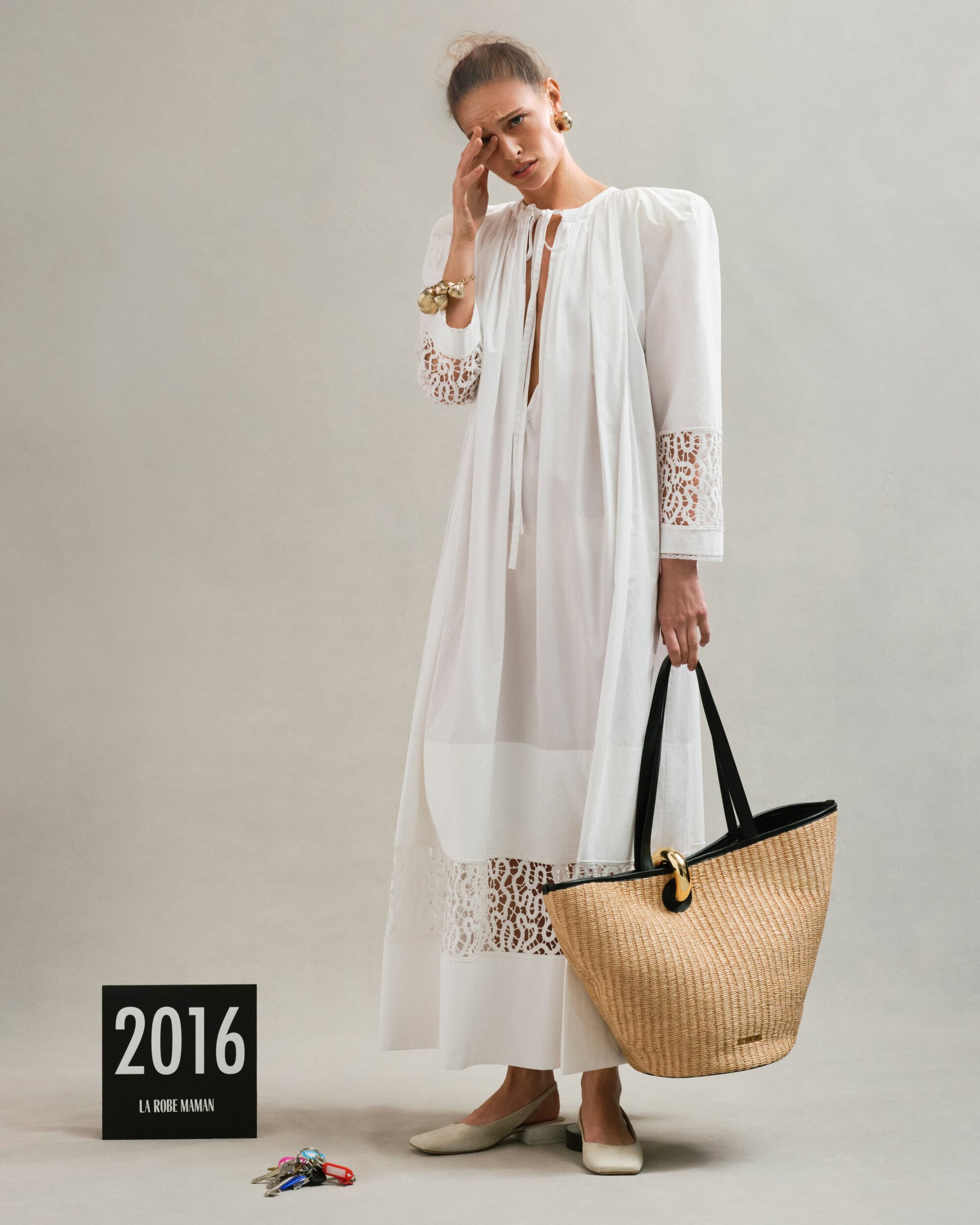 Model in long white lace-trimmed dress holding straw tote bag against a neutral studio backdrop for Jacquemus Spring 2025 Ad Campaign.