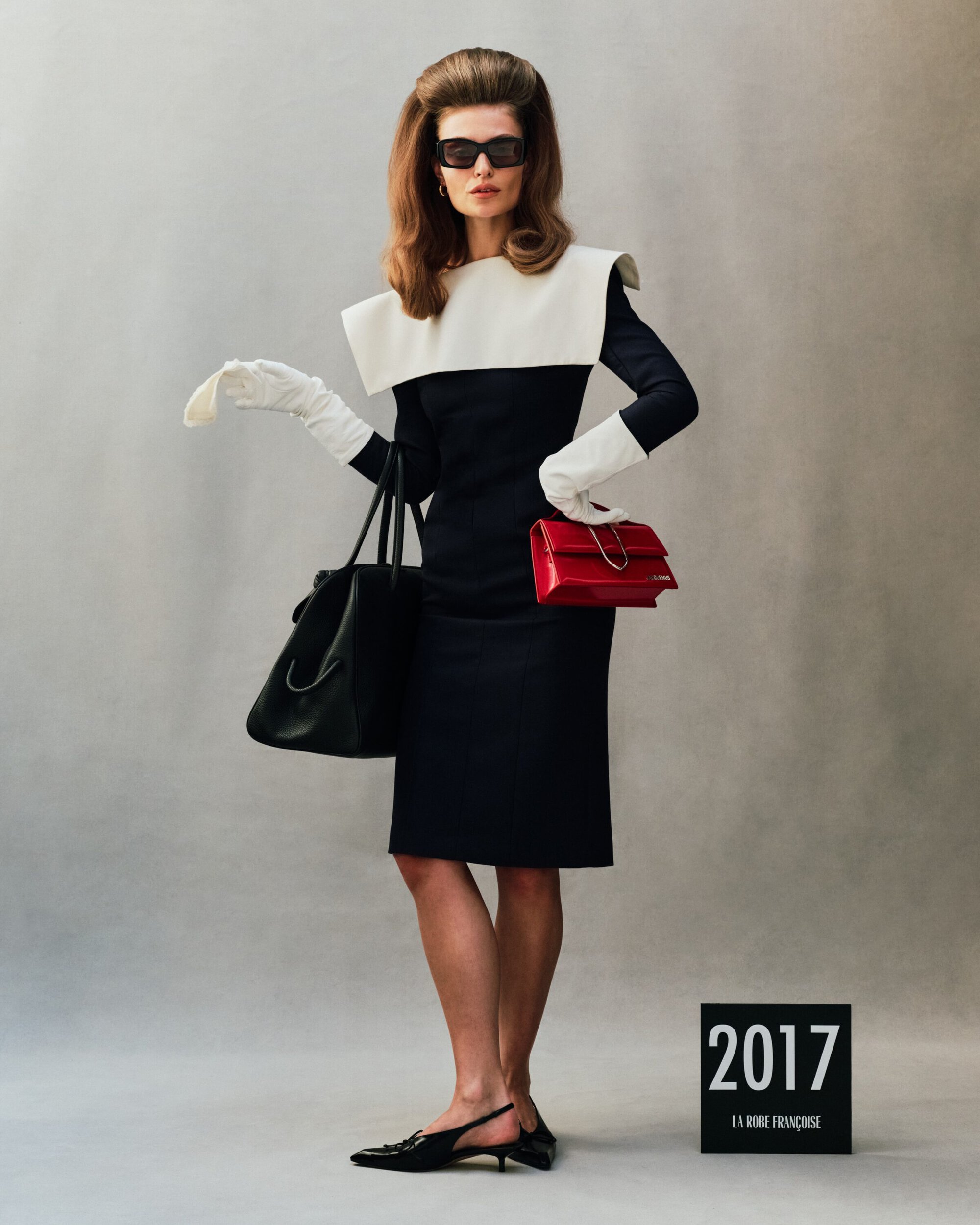 Model in dark sunglasses wearing a black and white dress, white gloves, holding a red handbag against a grey backdrop