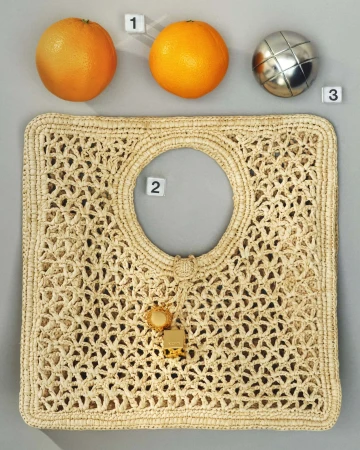 Overhead product flat lay of a woven straw handbag with two fresh oranges and a metallic sphere on grey seamless background