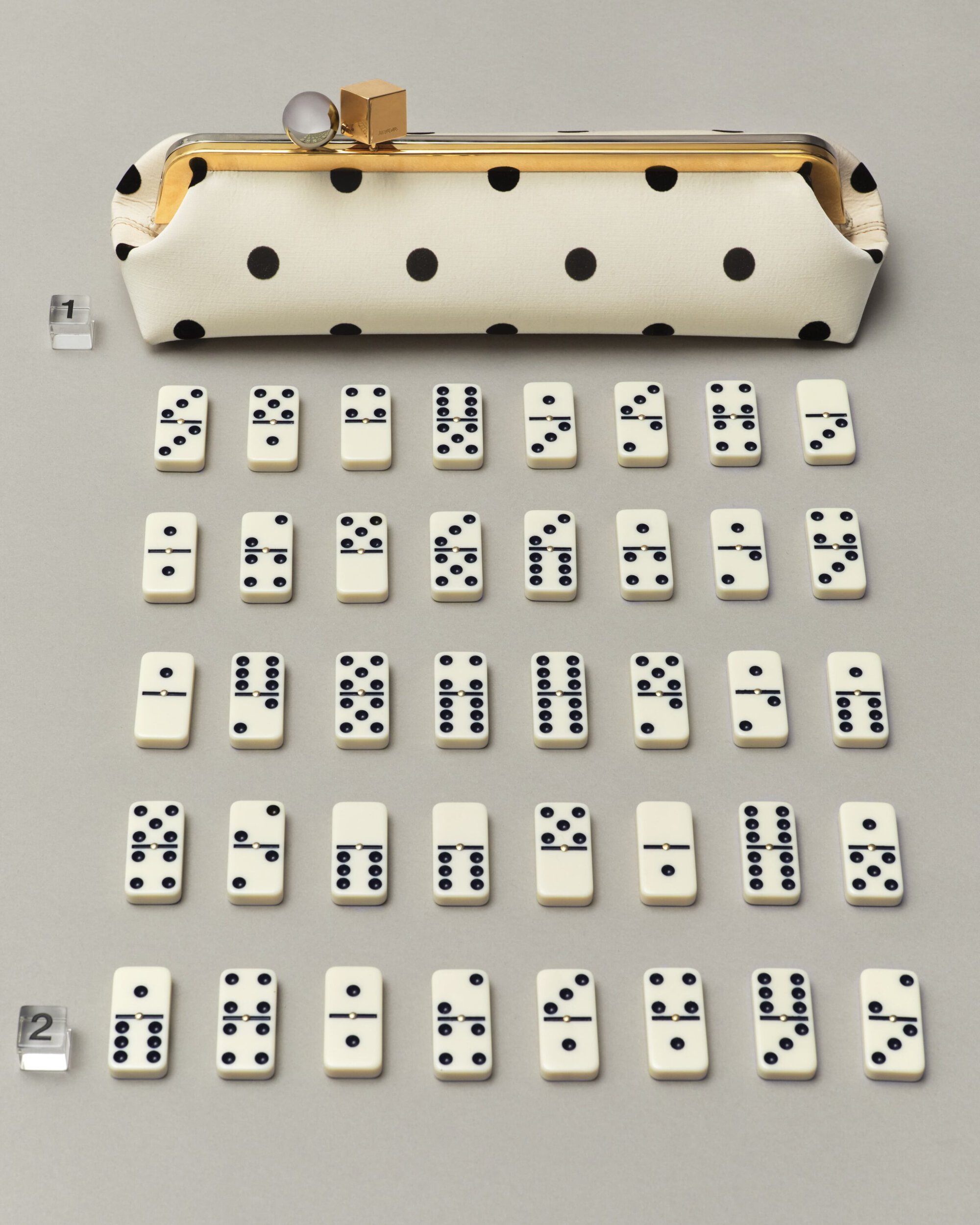 Polka dot pouch handbag with a gold clasp displayed above an arranged grid of dominoes on a grey seamless background.