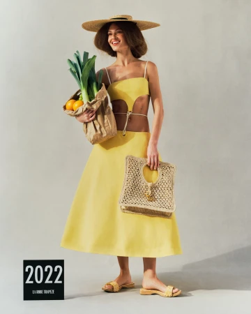 Model in a yellow cutout dress holding a paper grocery bag with leeks and oranges against a light grey seamless backdrop