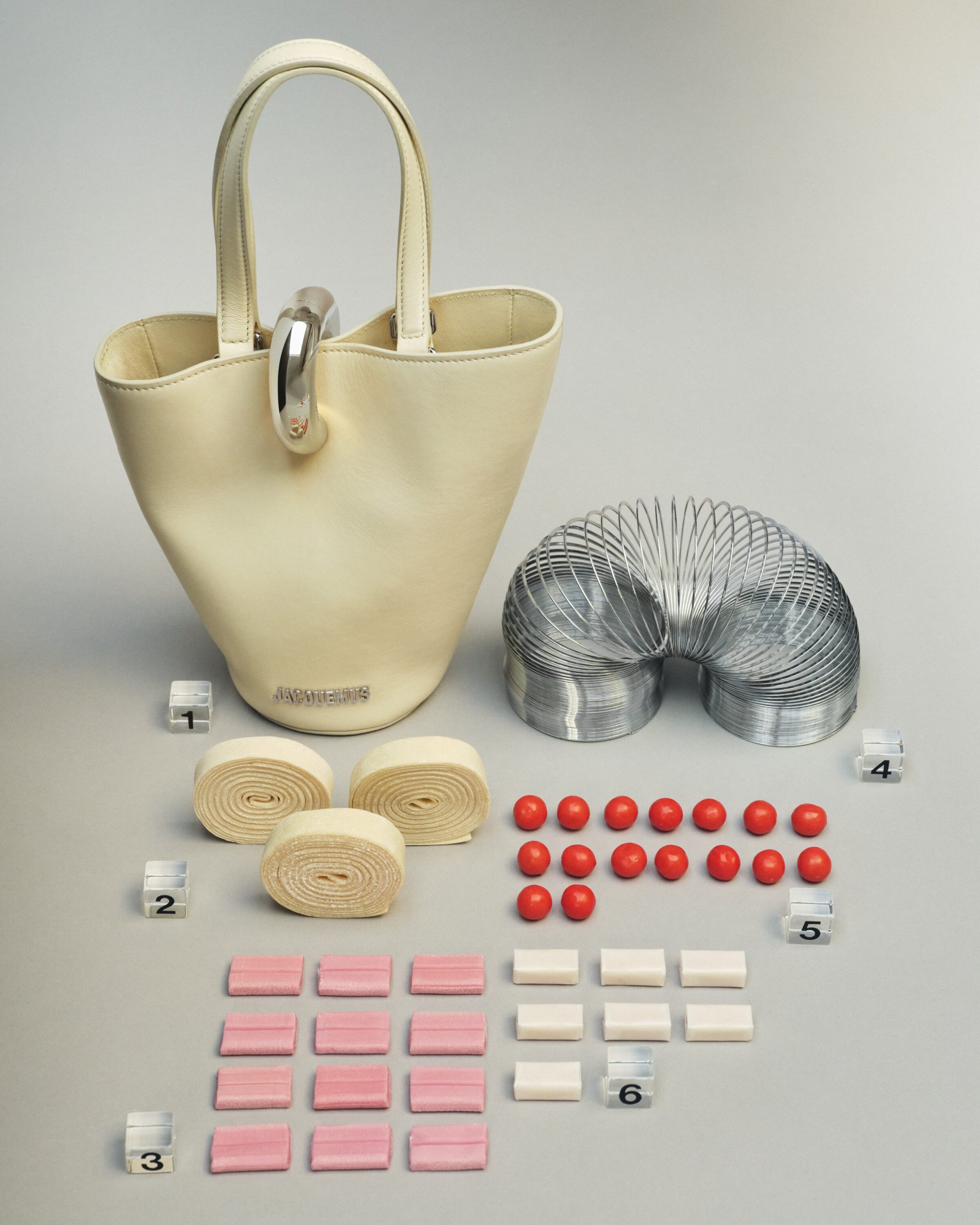 Cream leather bucket handbag with silver hardware displayed with a metal slinky, rolled ribbons, red spheres, and pink and white blocks.