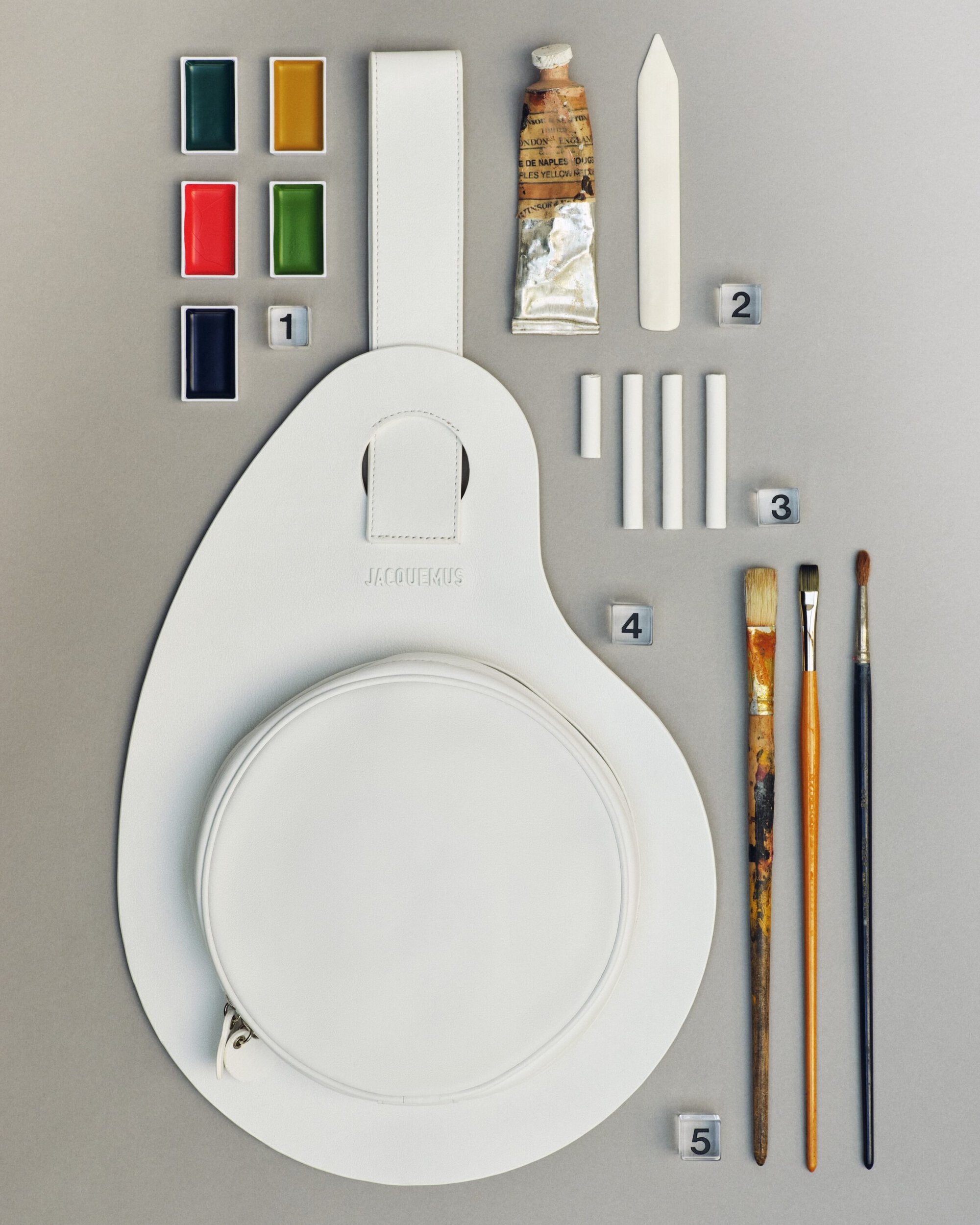 Flat lay of a white leather artist palette bag accompanied by paint pans, paint tube, chalk sticks, and brushes on grey