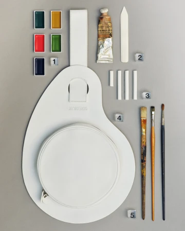 Flat lay of a white leather artist palette bag accompanied by paint pans, paint tube, chalk sticks, and brushes on grey