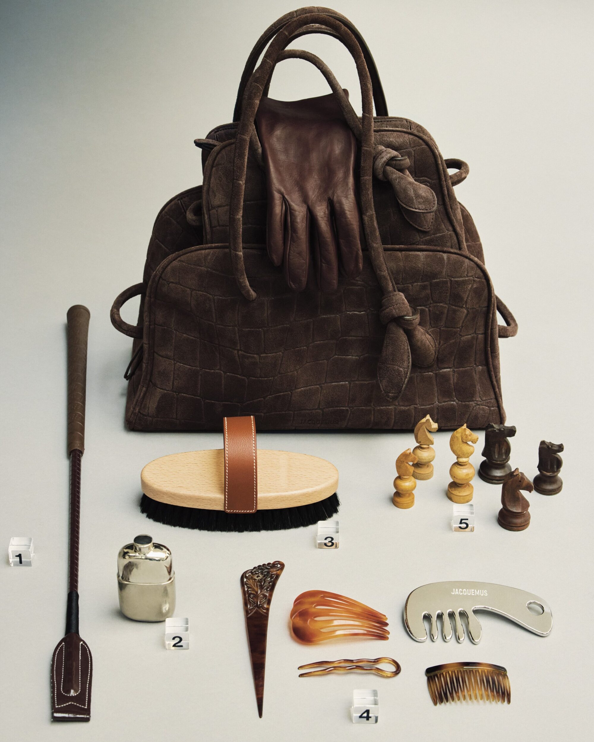 Brown leather handbag with gloves, riding crop, brush, chess pieces, and combs arranged on a neutral background.