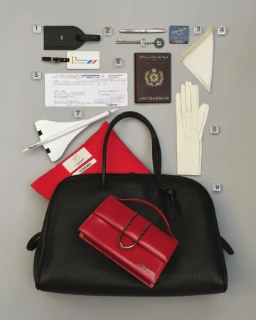 Flat lay of Jacquemus travel accessories including a black leather handbag, red handbag, passport, gloves, and airplane ticket on grey background