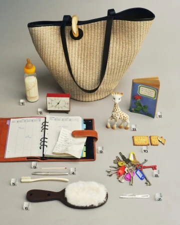 A straw tote bag with black handles and various everyday objects arranged on a neutral grey background.