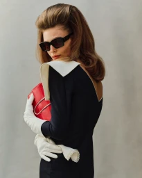 Woman wearing sunglasses, a black dress with white collar, white gloves, and holding a red handbag against a neutral wall