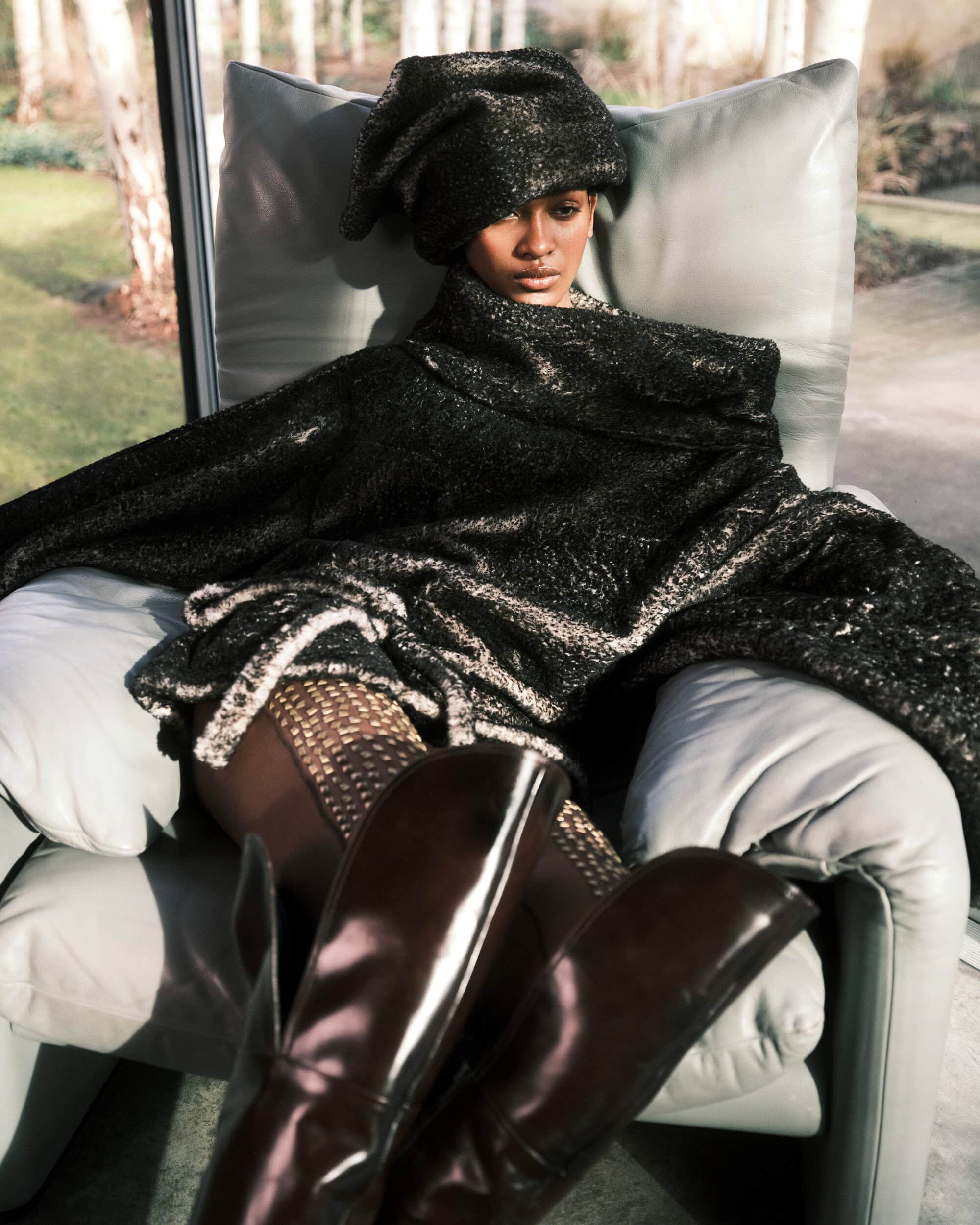 Model seated in an armchair wearing a dark textured oversized wrap and matching headpiece with tall boots.