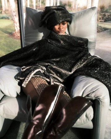Model seated in an armchair wearing a dark textured oversized wrap and matching headpiece with tall boots.