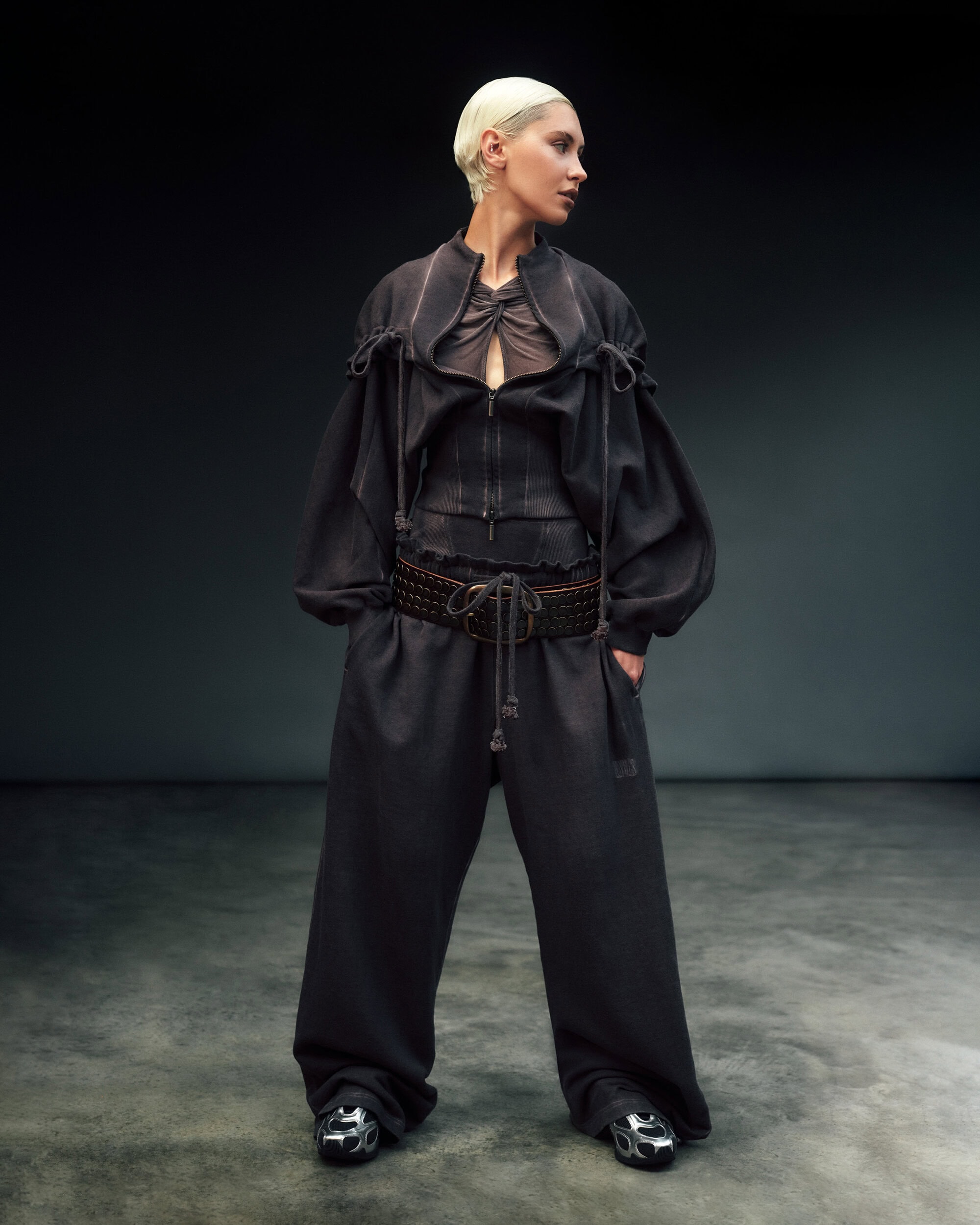 Model with blonde hair wearing dark grey voluminous outfit and studded belt against dark studio backdrop