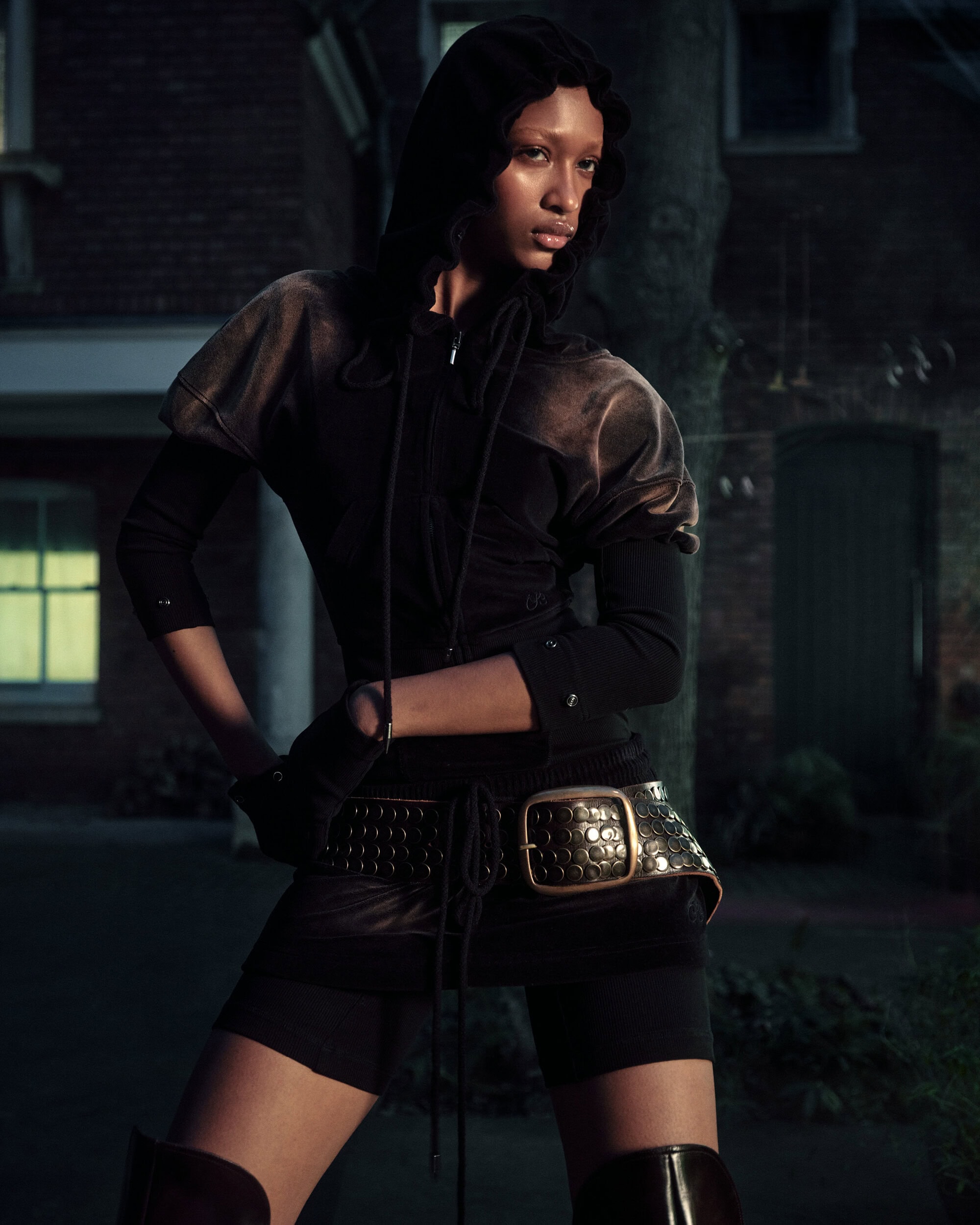 Model wearing dark hooded top and studded belt posing outdoors at dusk for KNWLS campaign