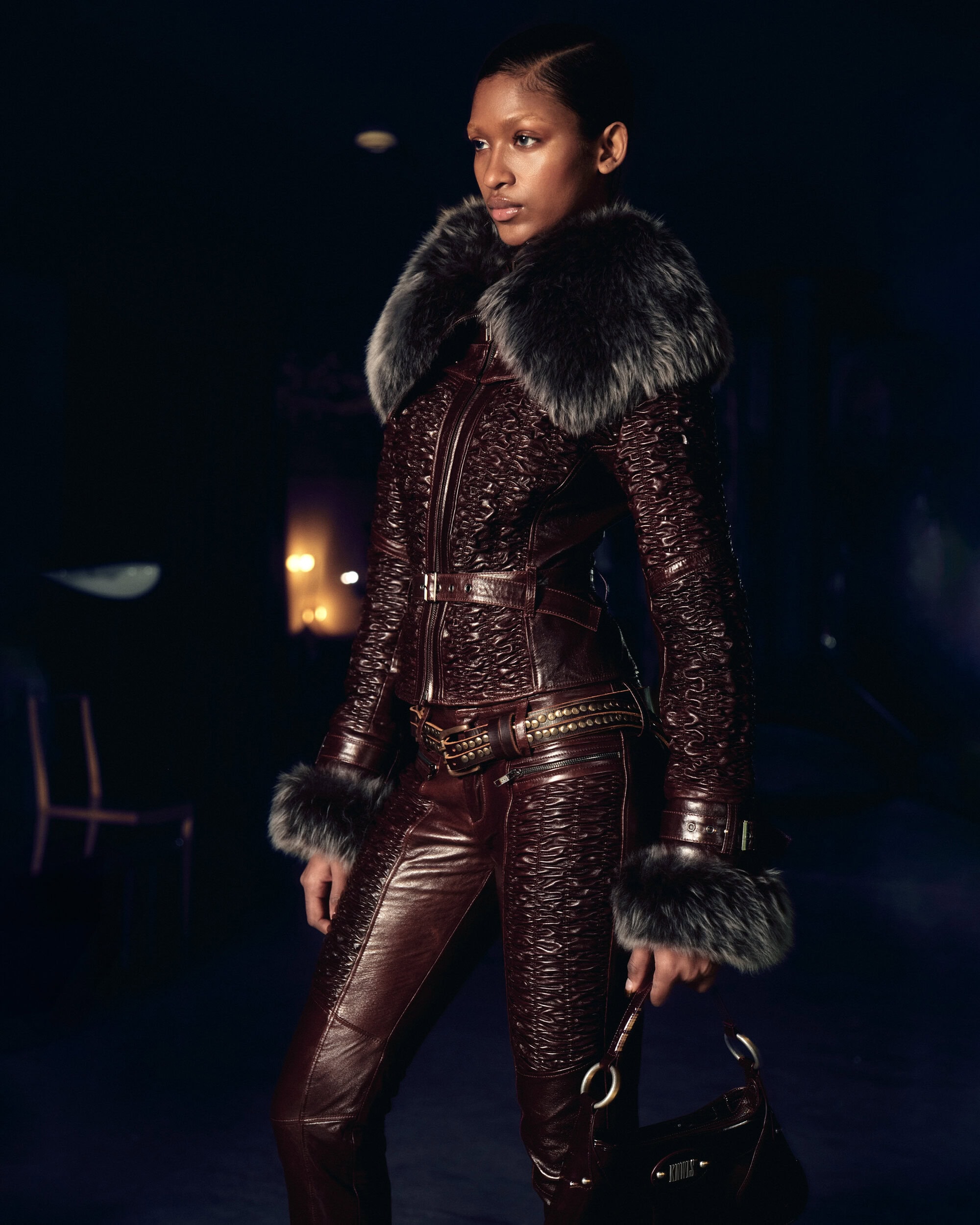 Model wearing a dark textured leather jacket with fur collar and matching trousers against a dark background