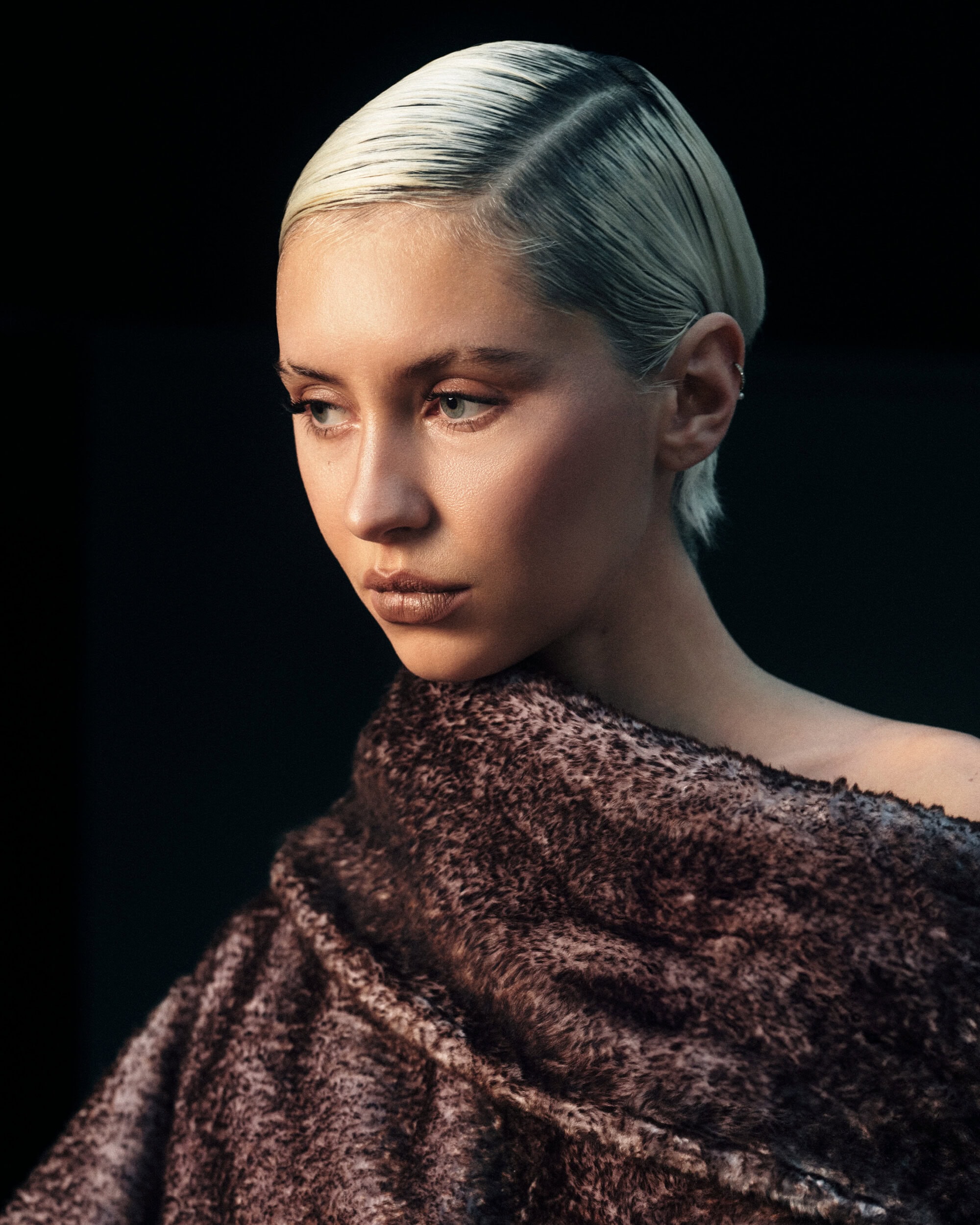 Portrait of a blonde woman wearing a textured brown draped garment against a dark background