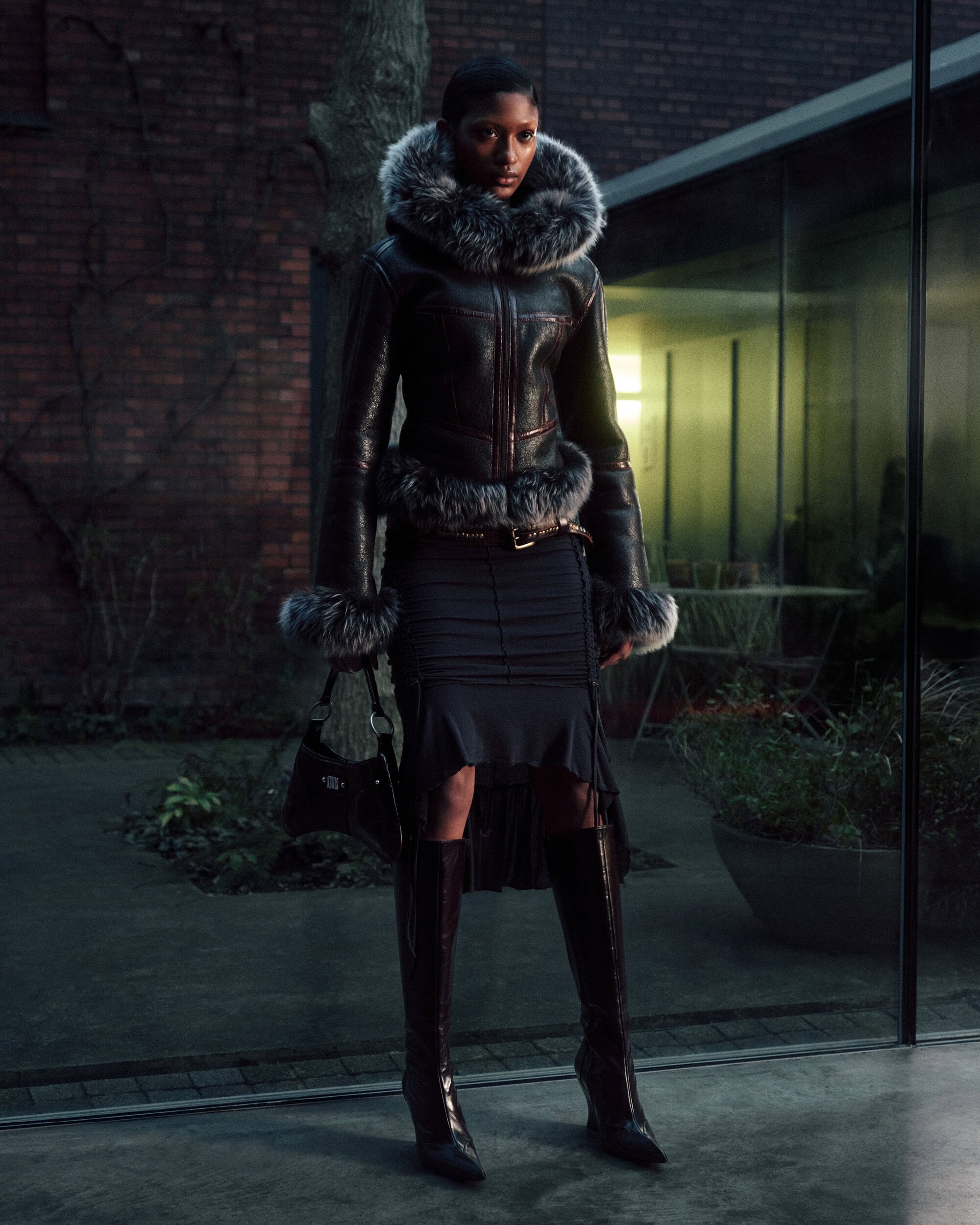 Model in a black fur-trimmed jacket and skirt standing outdoors near a brick building at dusk.