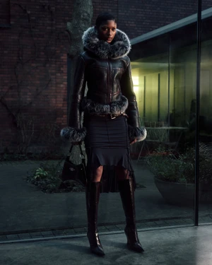 Model in a black fur-trimmed jacket and skirt standing outdoors near a brick building at dusk.