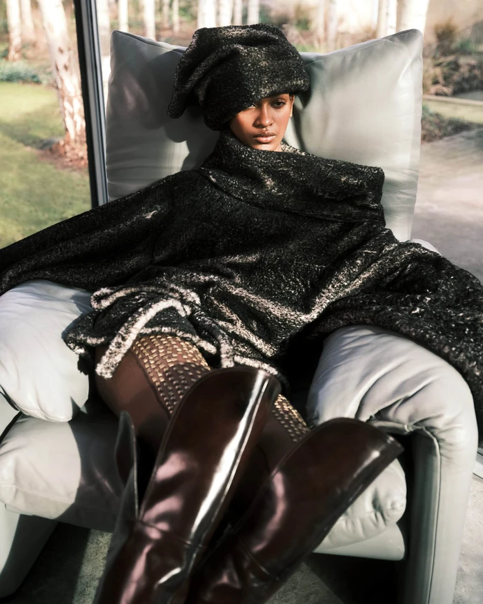 Model seated in an armchair wearing a dark textured oversized wrap and matching headpiece with tall boots.