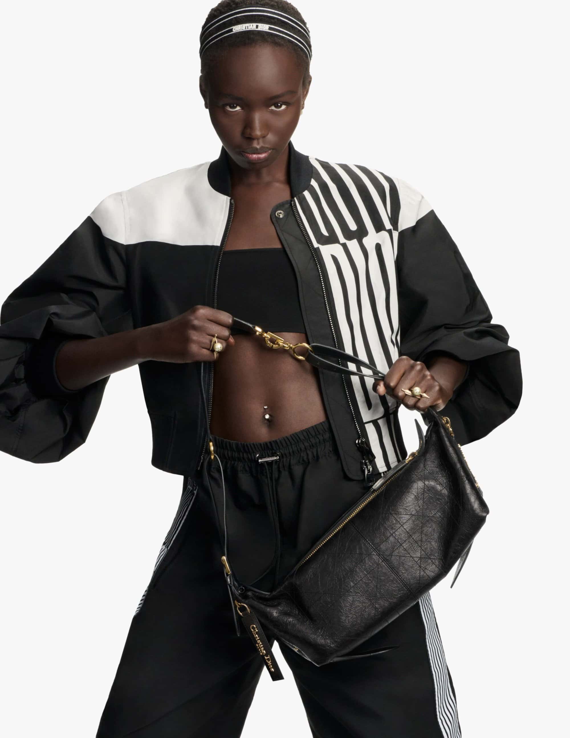 Model wearing a black and white bomber jacket holding a black leather handbag against a white background