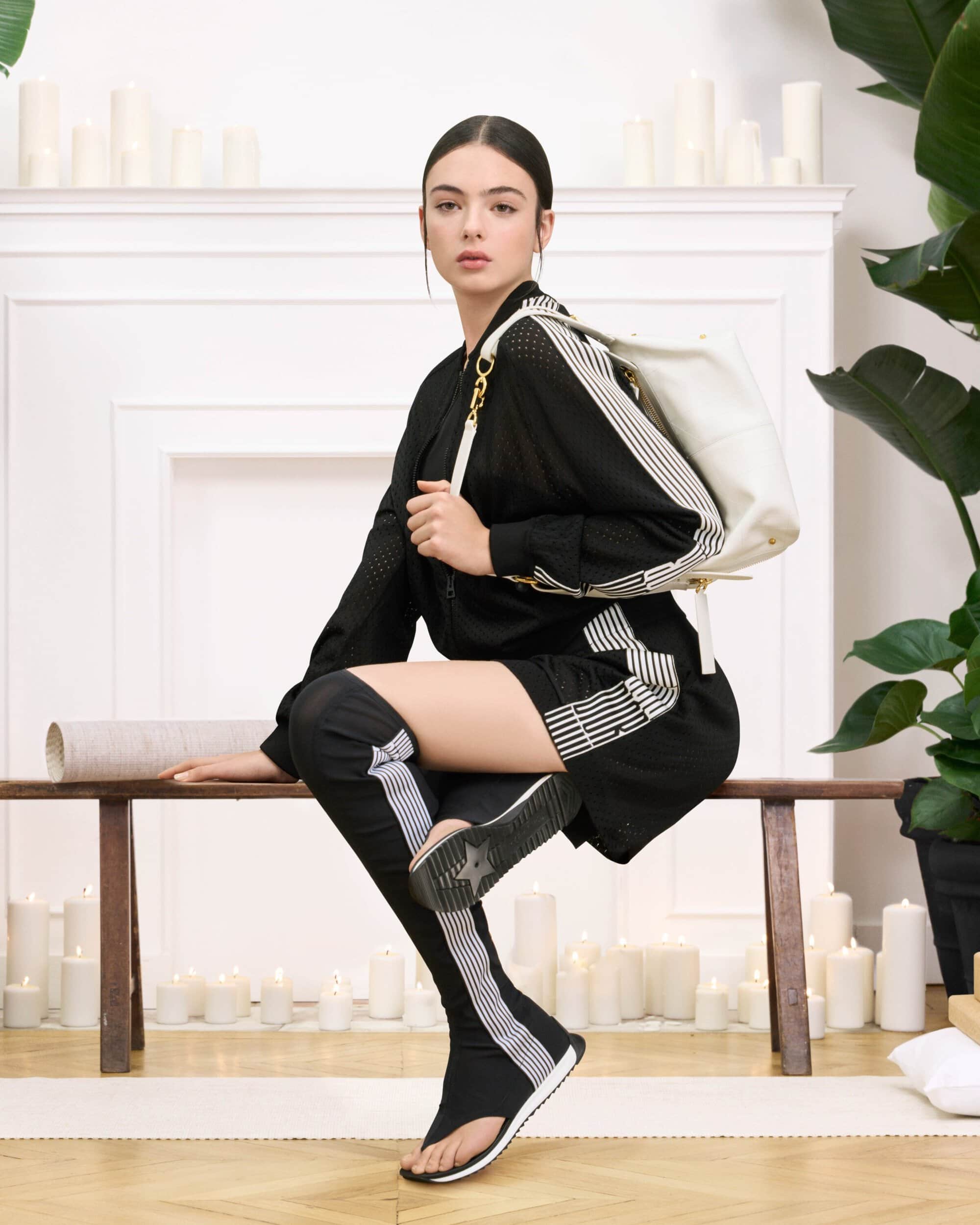 Model Deva Cassel posing on a wooden bench wearing a black athletic outfit with white stripes for Dior