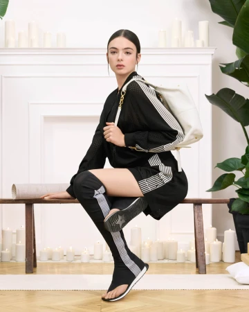 Model Deva Cassel posing on a wooden bench wearing a black athletic outfit with white stripes for Dior