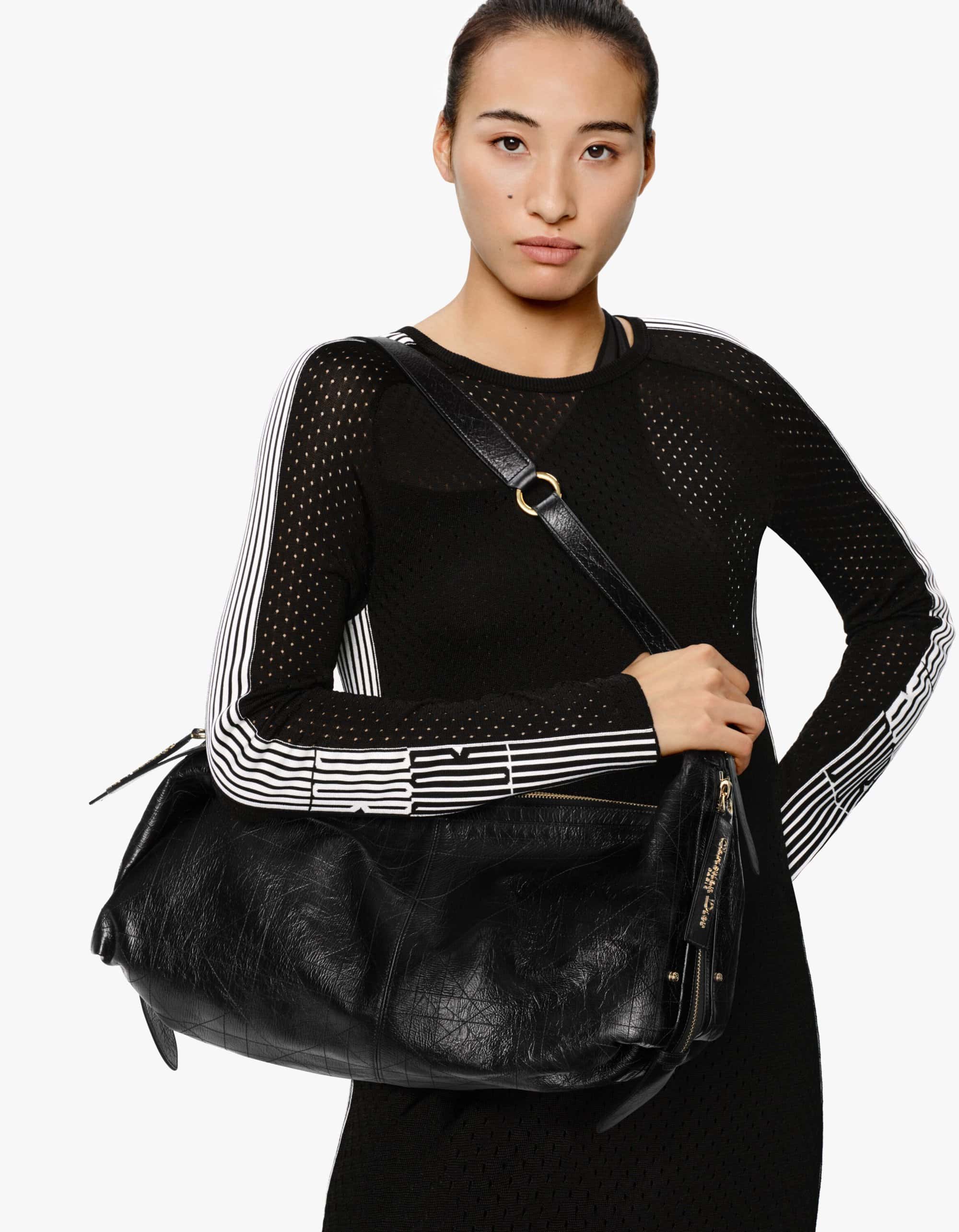 Model wearing black top and holding black leather shoulder handbag against a white background
