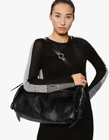 Model wearing black top and holding black leather shoulder handbag against a white background