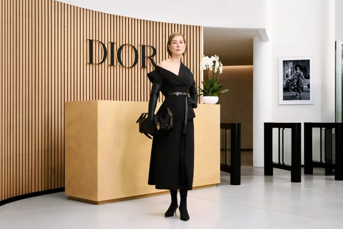 Rosamund Pike in a black off-shoulder dress and leather gloves holding a black handbag in a modern Dior interior