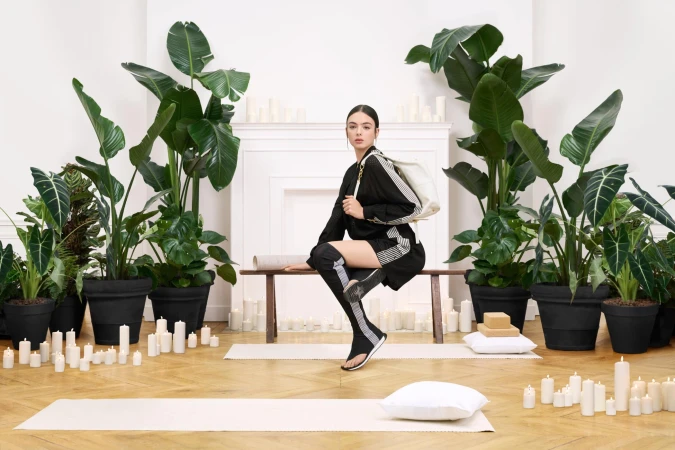 Model seated on a bench wearing a black outfit with white stripes, surrounded by potted plants and white candles