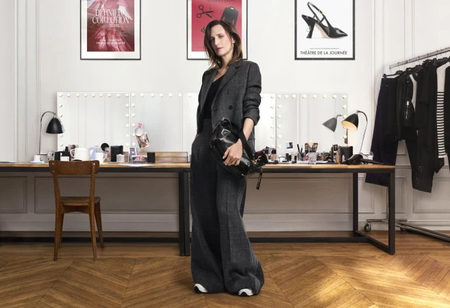 Camille Cottin wearing a dark grey suit holding a black handbag in a backstage dressing room
