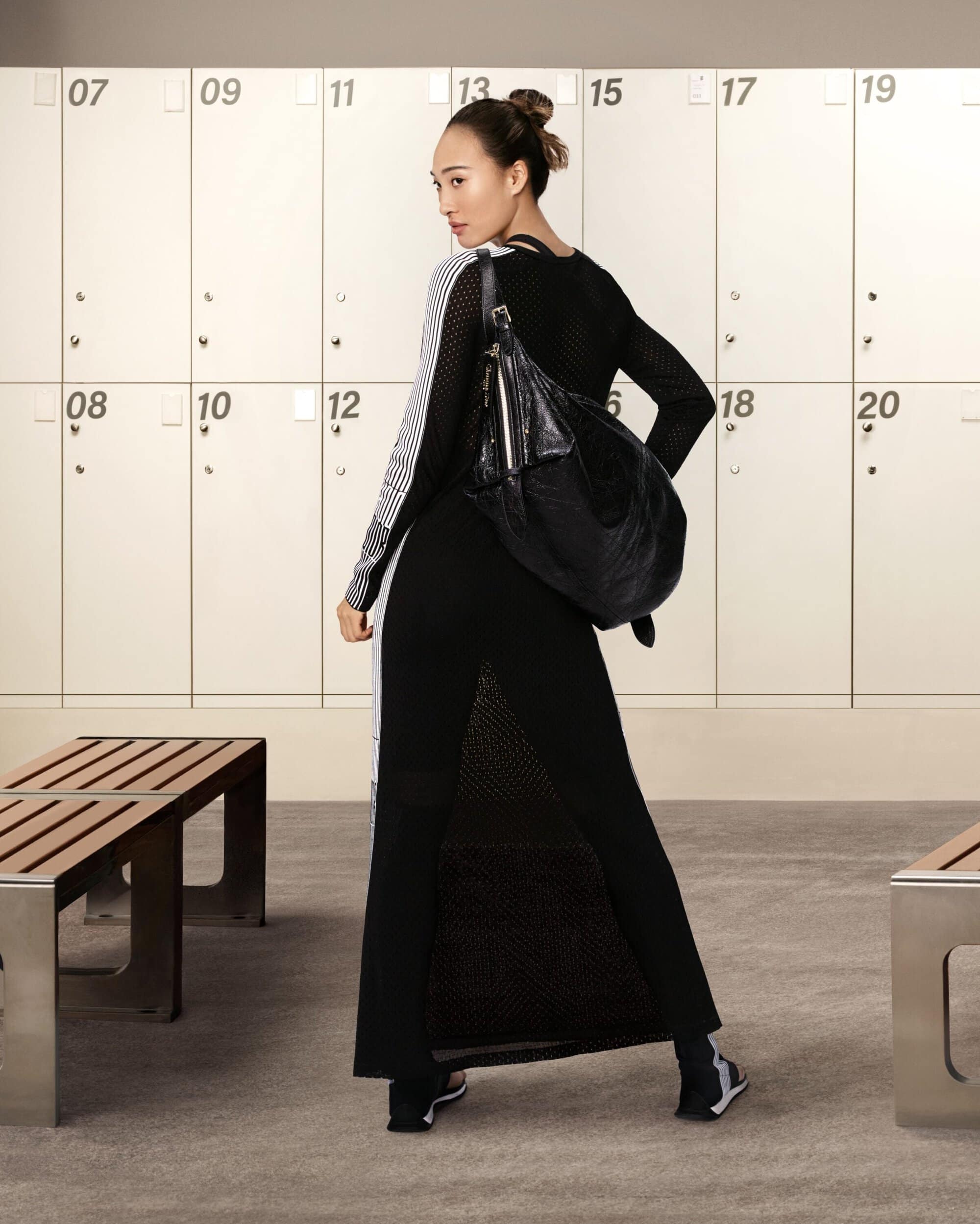 Woman in a black dress carrying a large black shoulder bag standing in front of lockers for Dior D-Journey campaign
