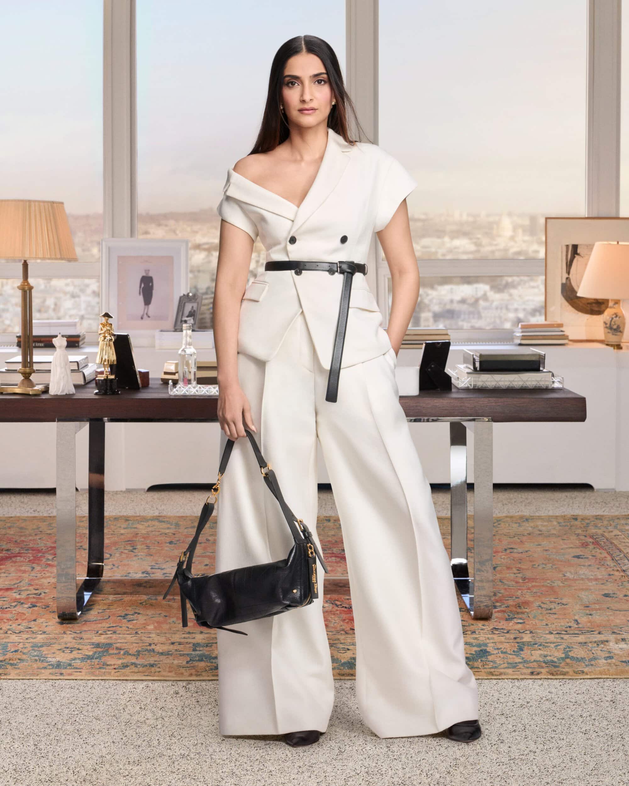 Sonam Kapoor wearing an off-shoulder white suit holding a black handbag in an elegant room interior