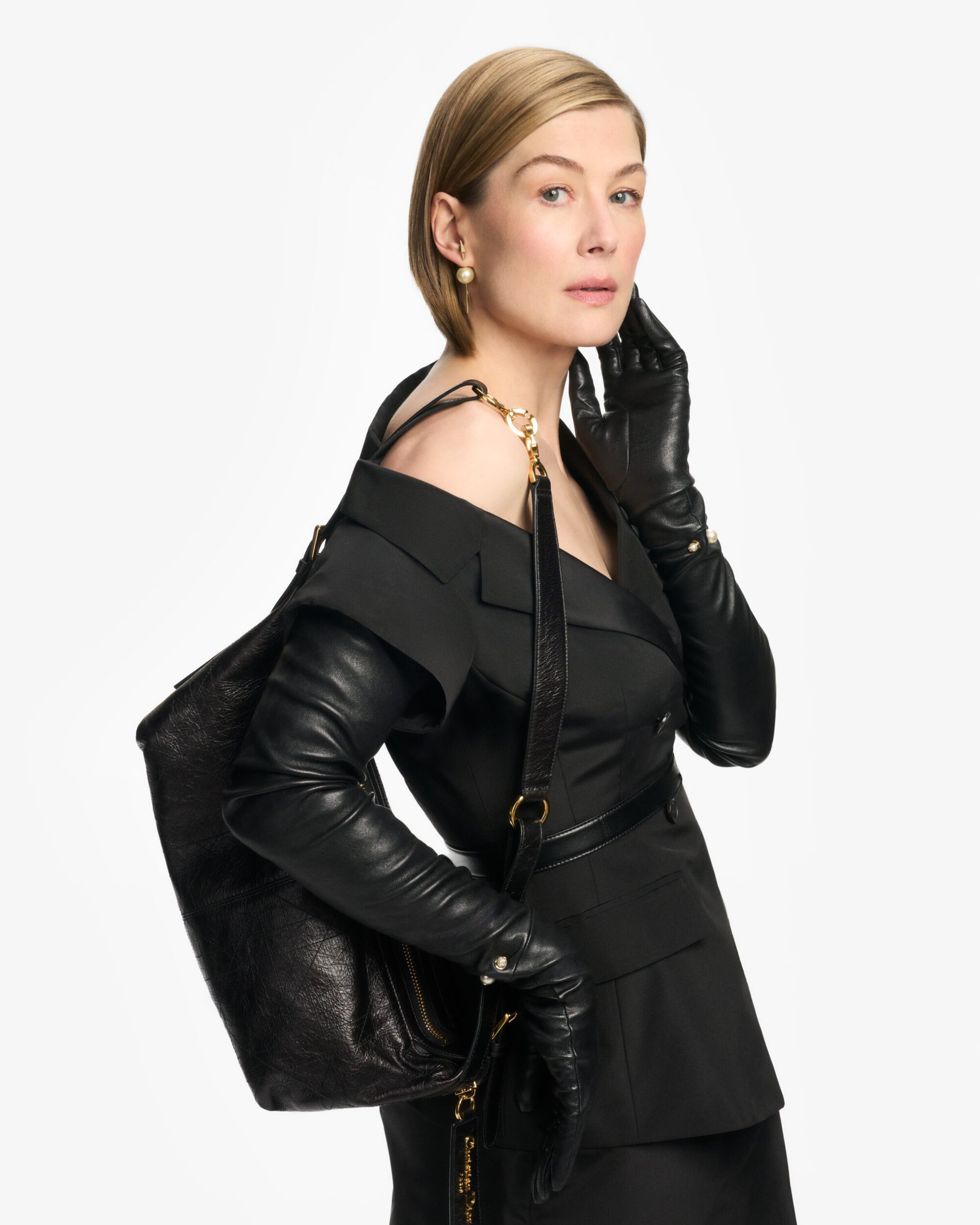 Rosamund Pike wearing a black dress and leather gloves carrying a black handbag against a white background for Dior