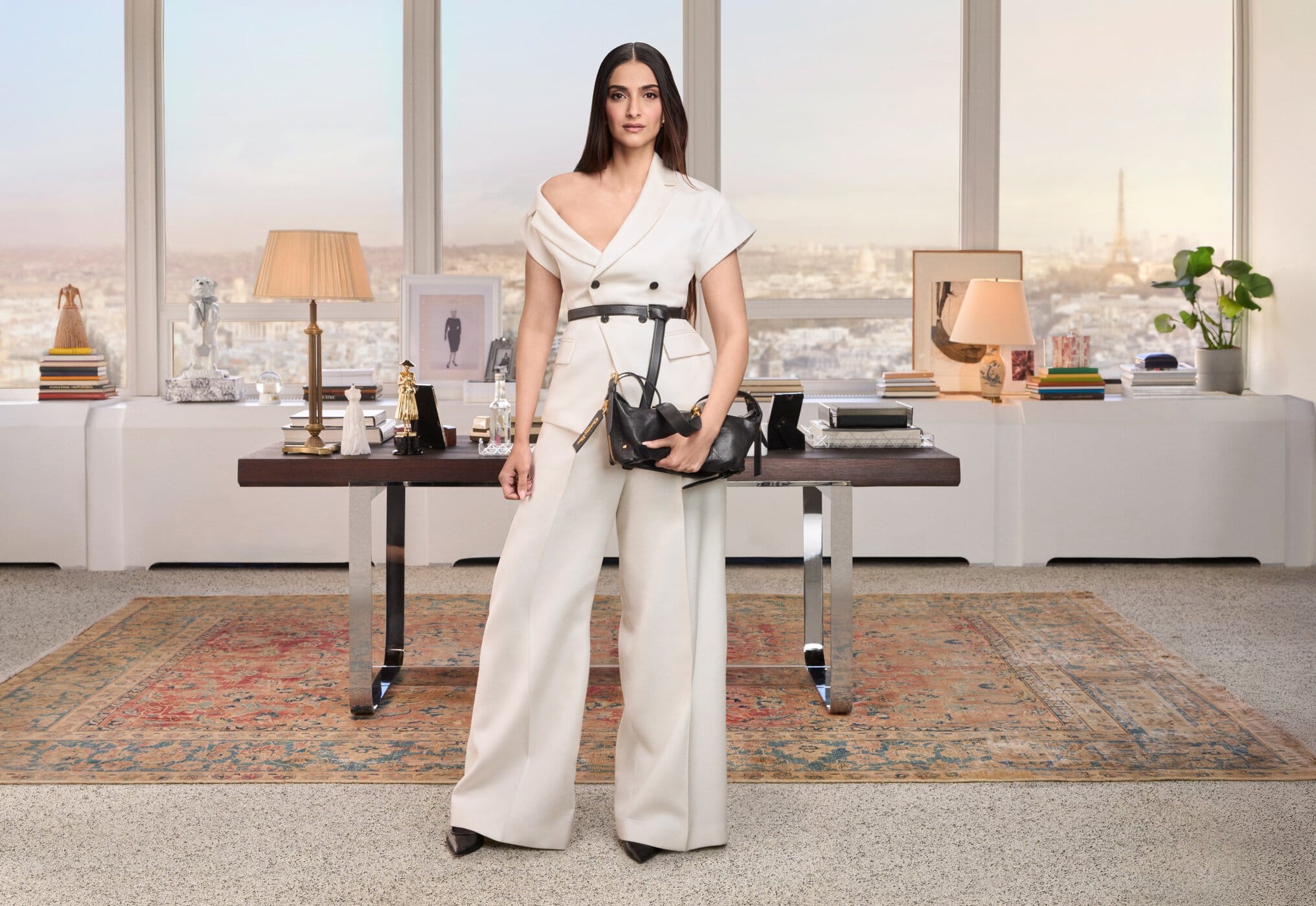 Sonam Kapoor in an off-the-shoulder cream pantsuit holding a black handbag in an office setting with large windows overlooking a city.