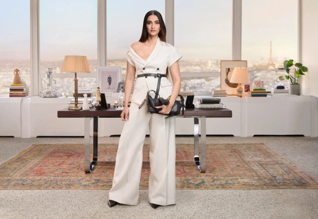 Sonam Kapoor in an off-the-shoulder cream pantsuit holding a black handbag in an office setting with large windows overlooking a city.