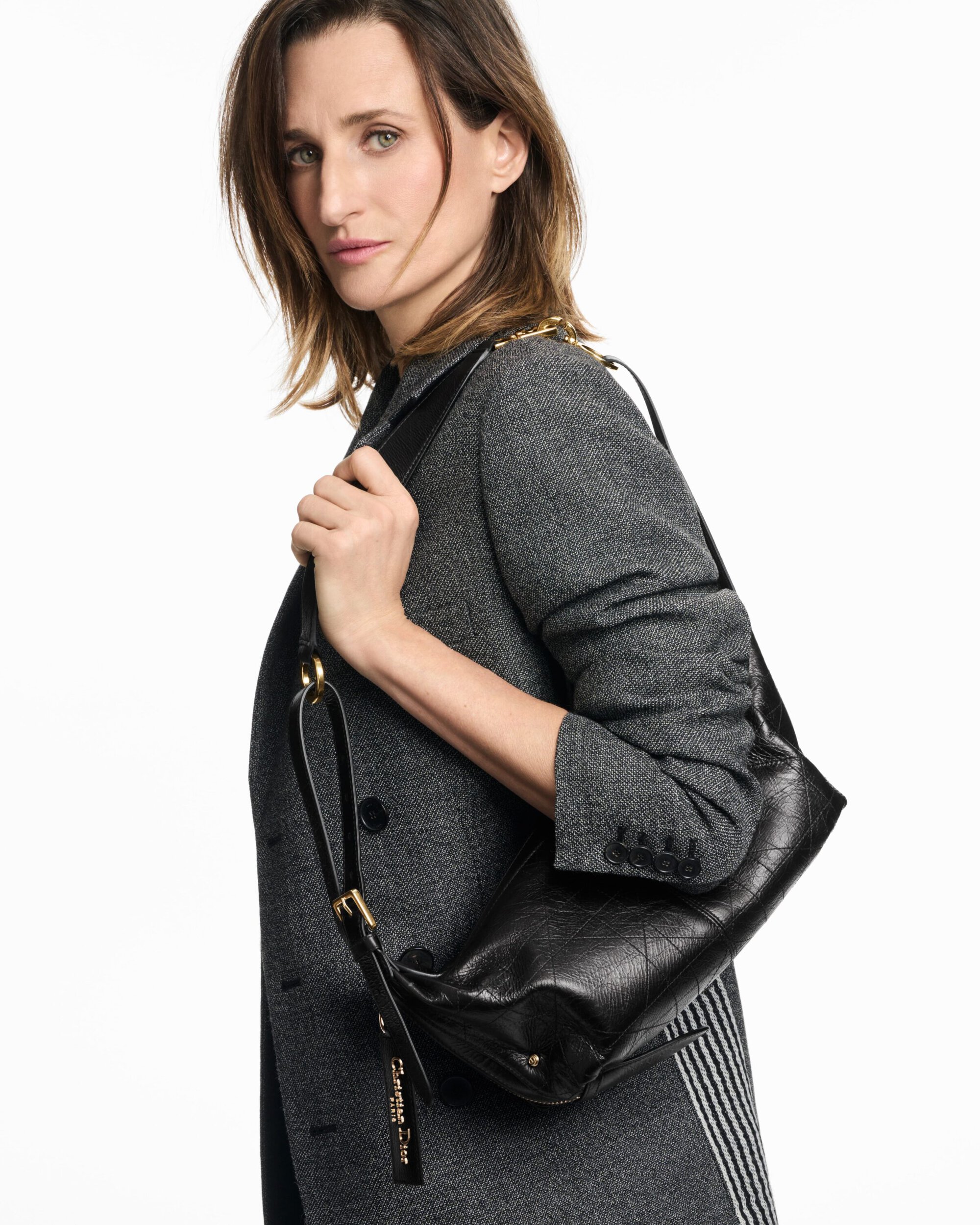 Camille Cottin wearing a grey coat and holding a black leather shoulder bag against a white background