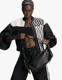 Model wearing a black and white bomber jacket holding a black leather handbag against a white background