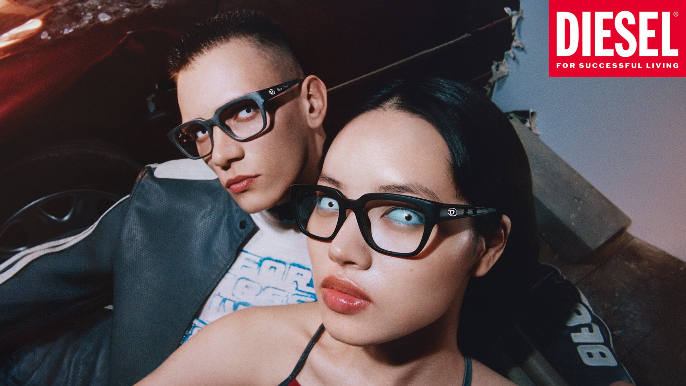 Two models wearing thick black-rimmed glasses and dark jackets in a dimly lit setting for Diesel Spring 2025 campaign.