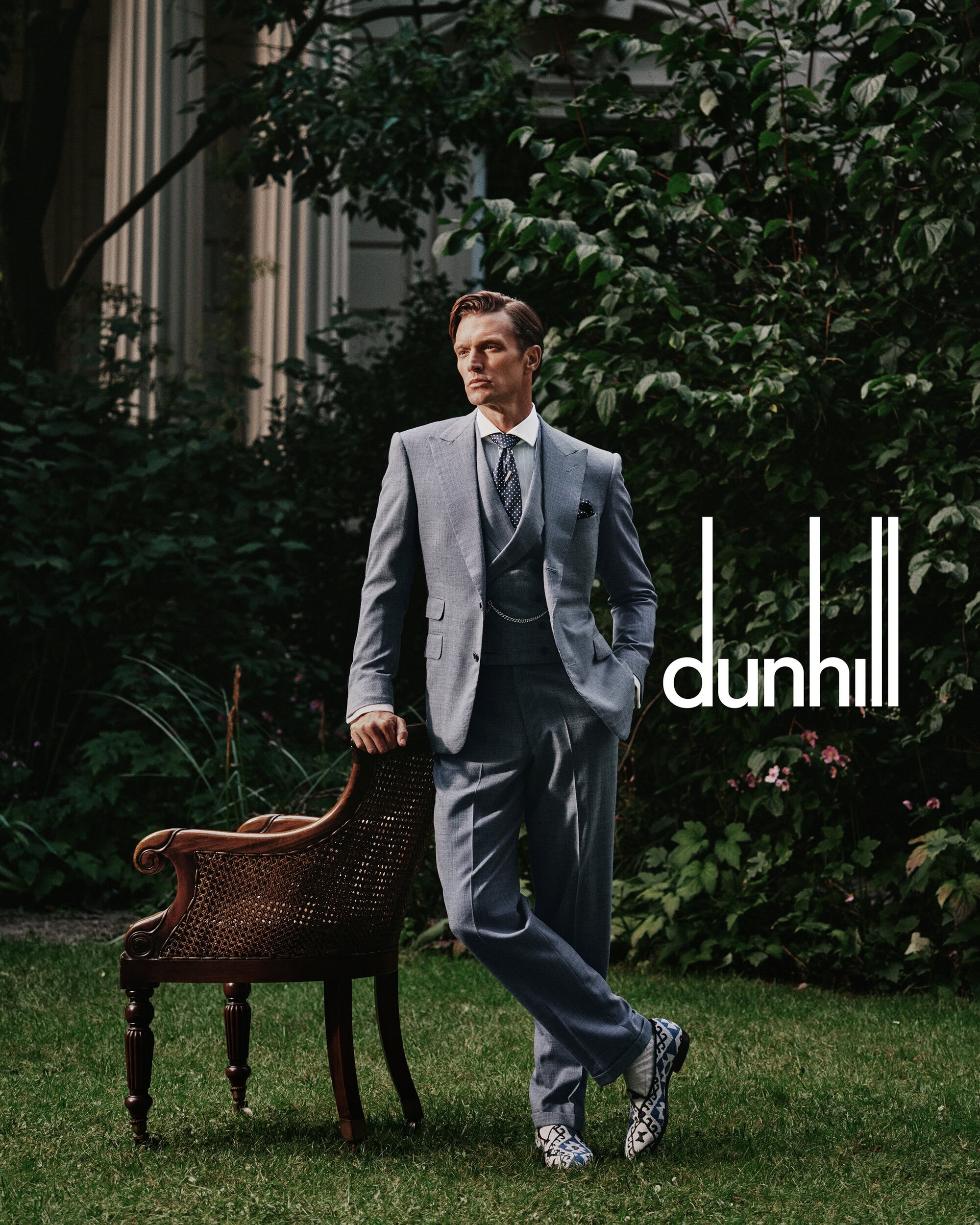 Man wearing a grey three-piece suit leaning against a vintage wooden chair outdoors in a garden