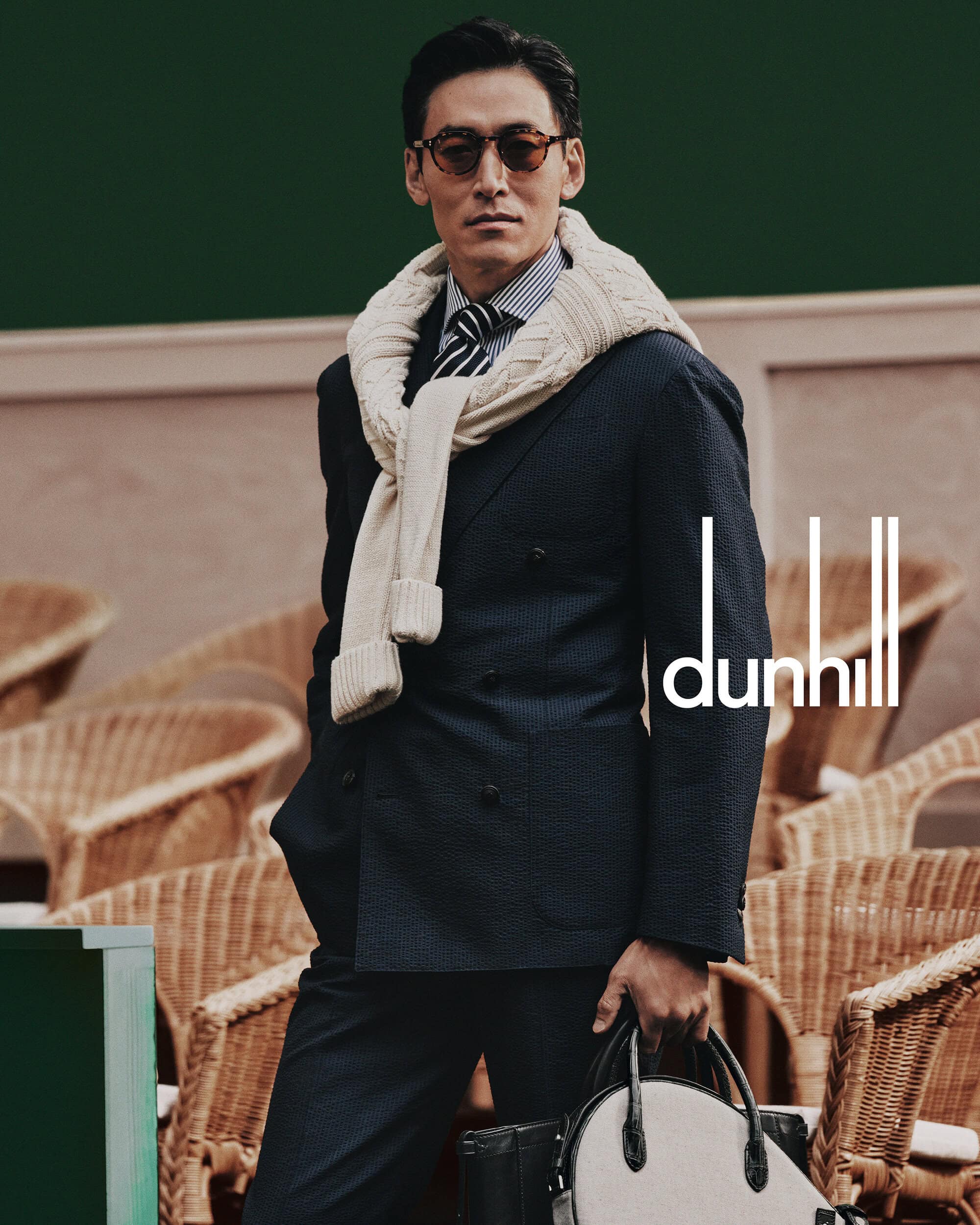 Man wearing a dark suit with a cream sweater tied over shoulders and holding a bag for Dunhill's Spring 2025 campaign.