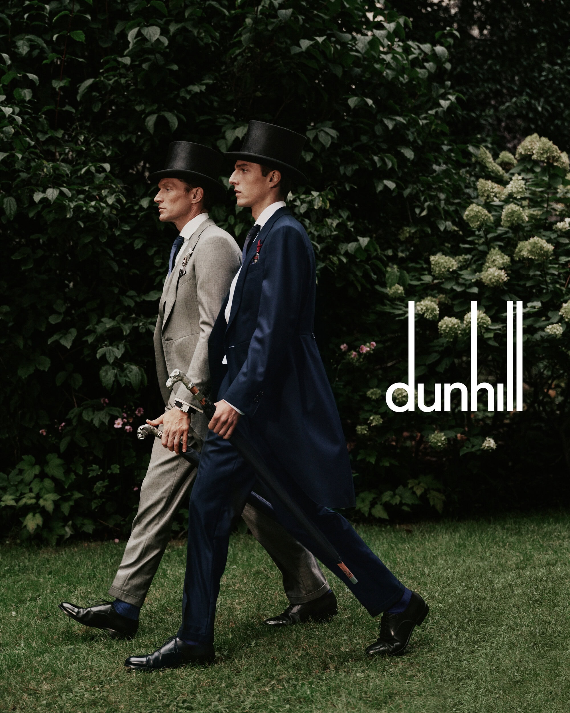 Two men in formal morning suits and top hats walking outdoors past green foliage for a Dunhill advertising campaign image.