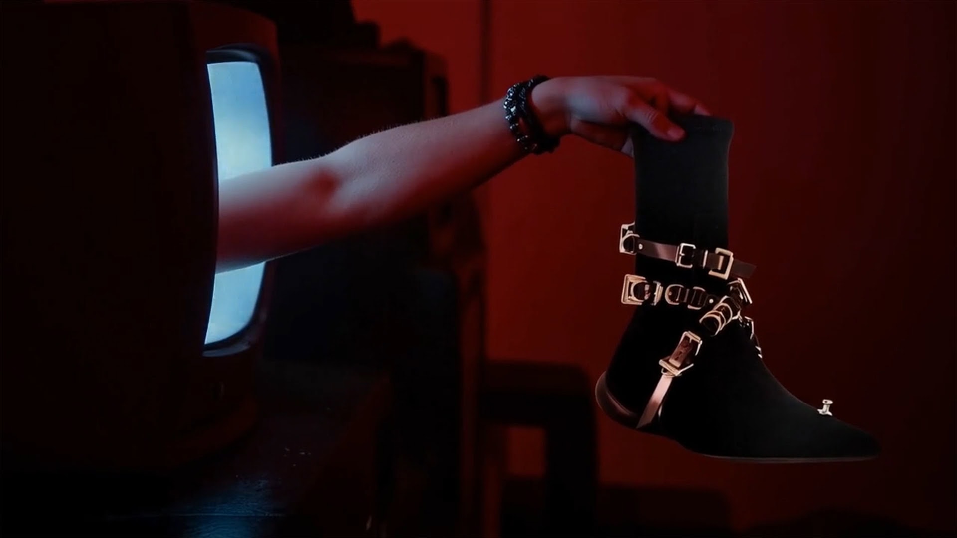 An arm reaches out of a glowing television screen to hold a black buckled ankle boot in a dimly lit red room.