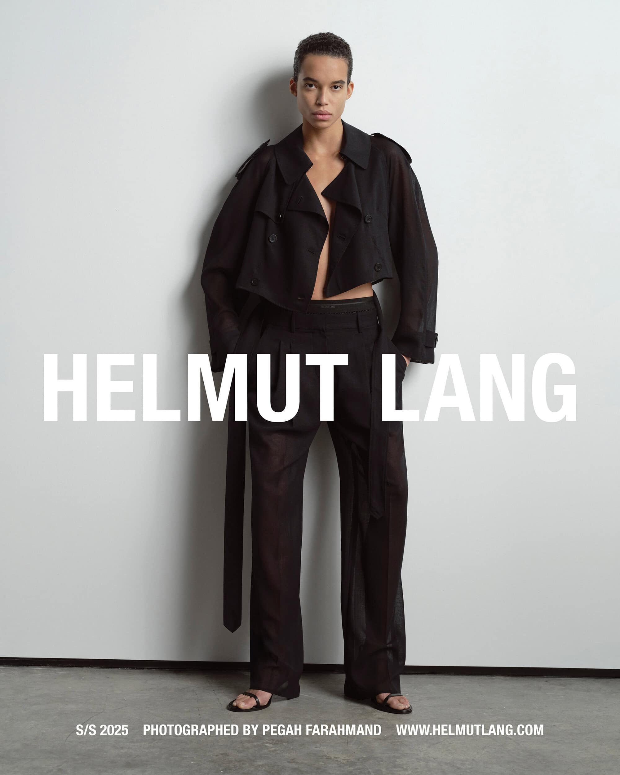 Model in black jacket and trousers standing against a pale grey studio wall for Helmut Lang
