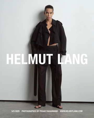 Model in black jacket and trousers standing against a pale grey studio wall for Helmut Lang