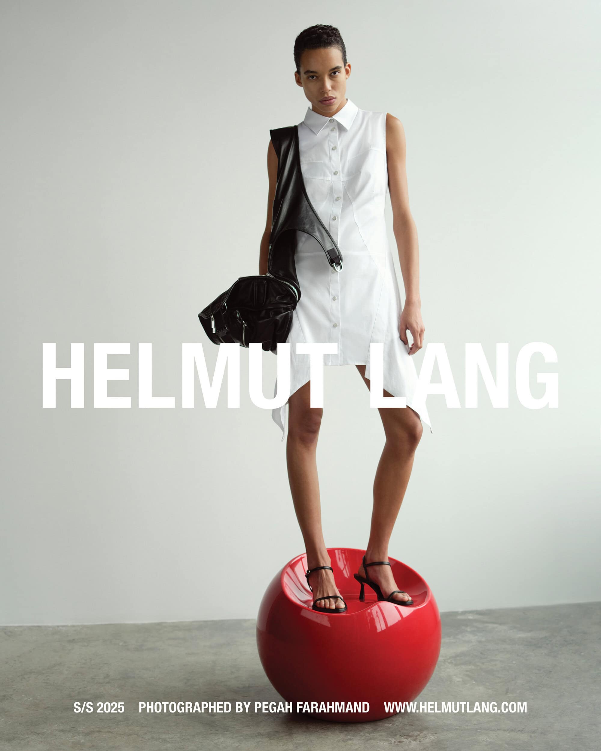 Model wearing a white sleeveless dress standing atop a red sphere holding a black bag against a plain studio background.