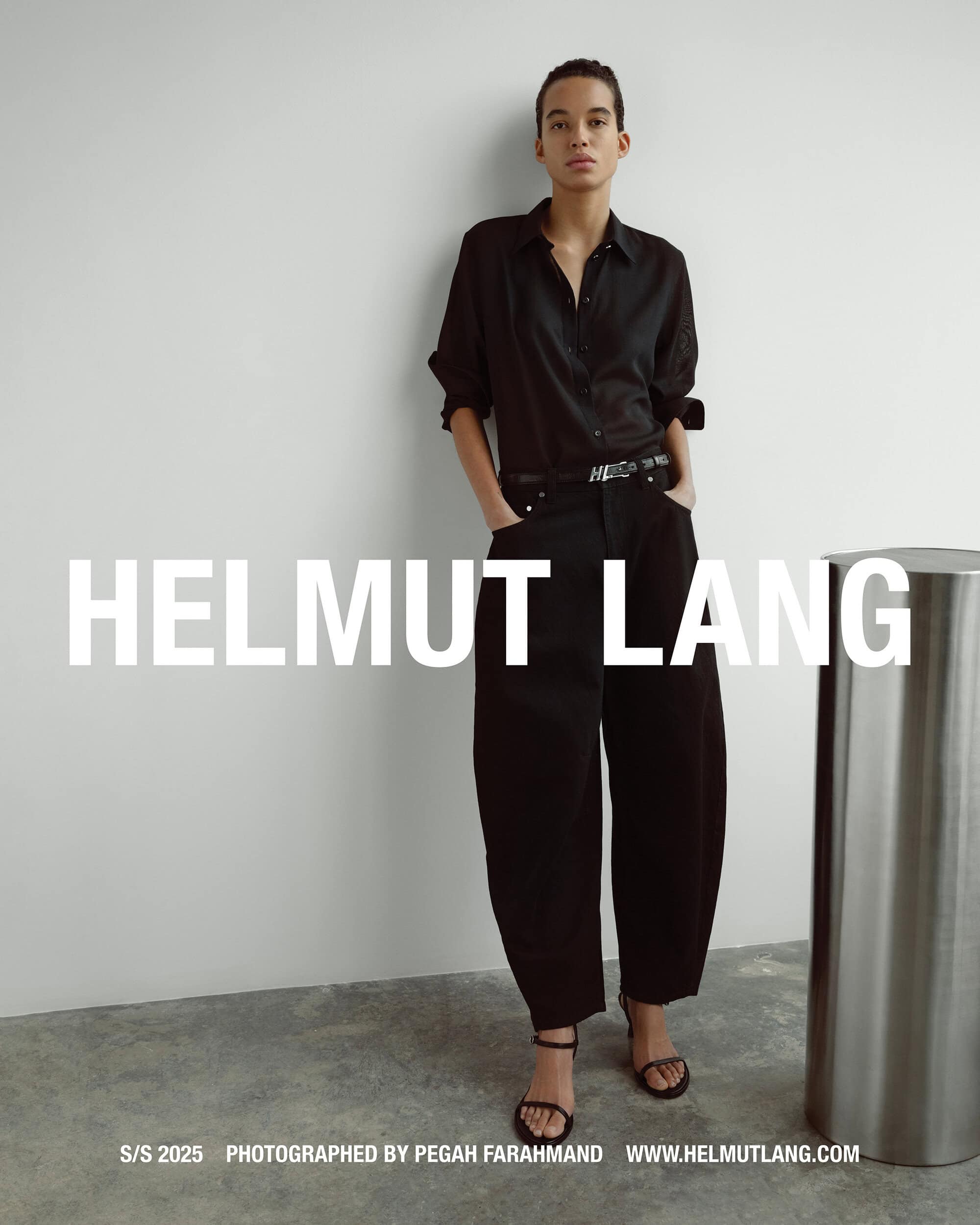 Model standing against a plain white wall wearing a black button-down shirt and black trousers with a metallic cylinder prop